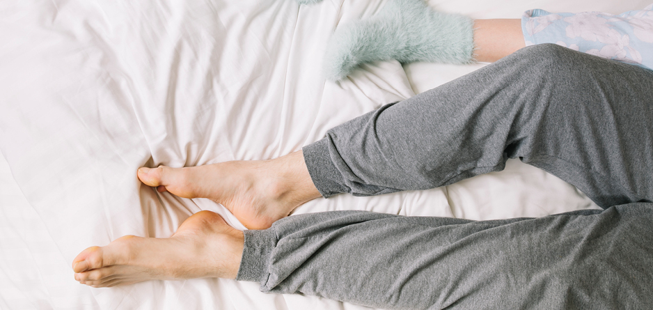 Two pairs of legs in bed, man in grey sweat pants and bare feet, woman in grey socks - leg cramps at nighttime causes and solutions.